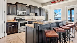 A modern kitchen with dark cabinetry, granite countertops, a large island with bar stools, and a view of the outdoors through large windows. - 7 acre Estate in Gated Reserve Community with Access to Pools, Beach, & Trails (Fish Haven)