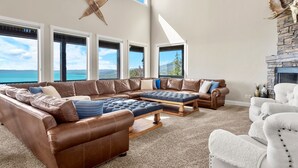 A spacious living room with a large brown leather sectional sofa, a white armchair, and a wooden coffee table. There are large windows offering a view of the outdoors. - 7 acre Estate in Gated Reserve Community with Access to Pools, Beach, & Trails (Fish Haven)