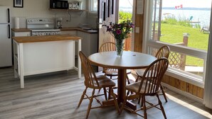 Een keuken met een kookplaat, magnetron en koelkast. Er is een eettafel met stoelen en een vaas met bloemen. Een raam biedt uitzicht op een meer en er staan stoelen buiten.