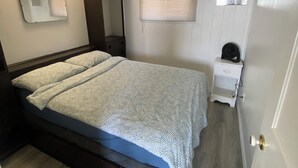 Een slaapkamer met een bed, nachtkastje, spiegel, raam met zonwering, en een deur.
