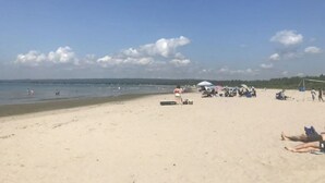 Une plage où l’on peut voir des gens se prélasser au soleil et nager, ainsi que des tentes, le tout sous un ciel dégagé.