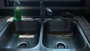 Een keuken met een roestvrijstalen spoelbak, een fles afwasmiddel en een raam met uitzicht op een grasveld met mensen en een waterpartij.