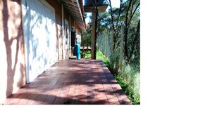 A wooden deck with a metal railing, surrounded by greenery and a fence. - Beautiful single-story house, super cozy: 3 suites. (Monte Verde)