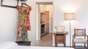 Habitación de hotel con una cama, una silla, una mesita con lámpara, y un vestido colorido colgado en la pared.