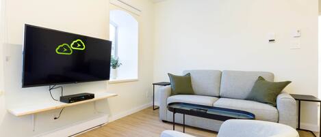 A modern living room with a flat-screen TV, a grey sofa, a small side table, and a light-colored armchair.