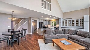 Ein geräumiges Wohnzimmer mit einem großen grauen Ecksofa, einem Holztisch und einem Essbereich mit rundem Tisch und Stühlen. Eine Treppe führt zu einer oberen Ebene mit einem Balkon.