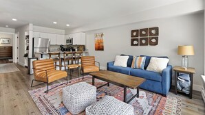 A modern living room with a blue sofa, wooden coffee table, and patterned ottomans. A kitchen with white cabinets and a dining area with wooden chairs is visible in the background. - The Valley Vista: Top floor unit w/ views & hot tub (Telluride)