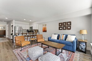 A modern living room with a blue sofa, wooden coffee table, and patterned ottomans. A kitchen with white cabinets and a dining area with wooden chairs is visible in the background.