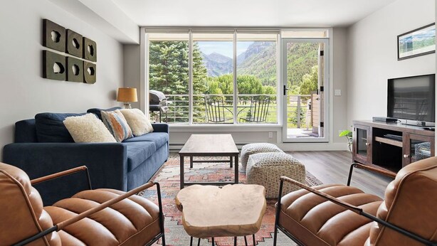A modern living room with a sofa, leather chairs, a coffee table, and a TV. A large window offers a view of mountains and greenery. - The Valley Vista: Top floor unit w/ views & hot tub (Telluride)