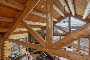 Ein Blockhaushaus-Innenraum mit hohen Decken, Holzbalken und großen Fenstern mit Blick auf die Berge.