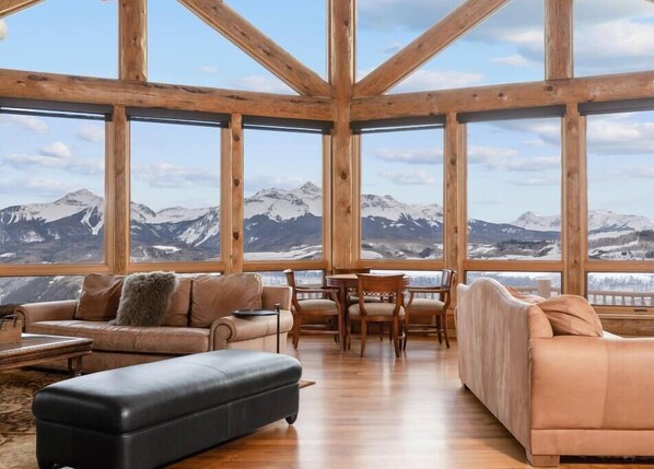 A spacious room with large windows offering mountain views, wooden beams, a leather sofa, a dining table with chairs, and a black ottoman. - 140 Serapio Ln (Telluride)