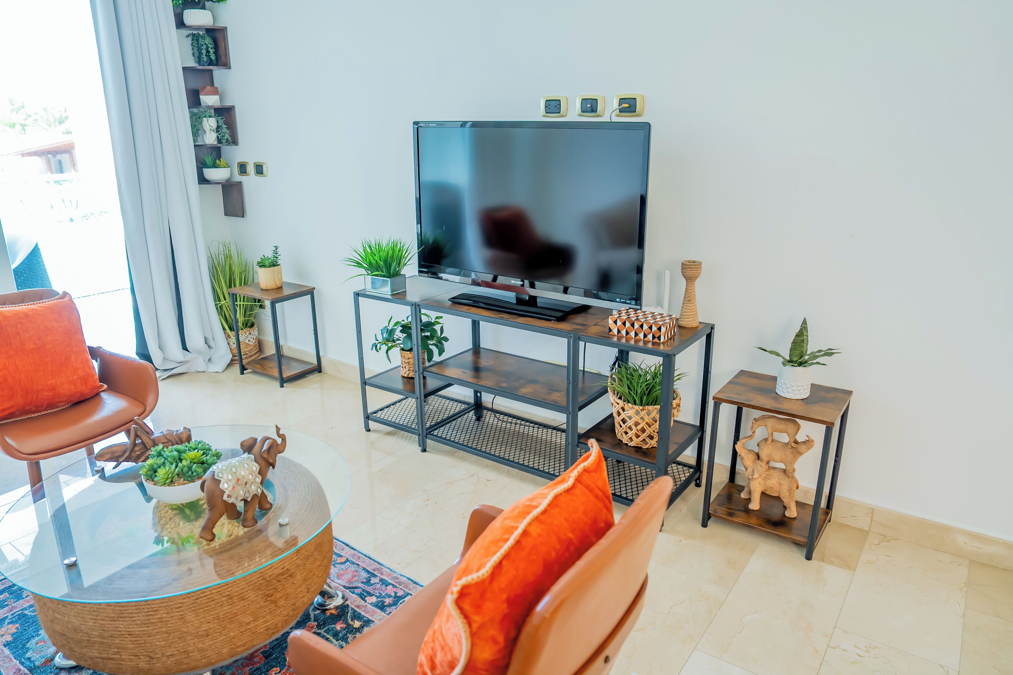 A modern living room with a glass coffee table, a comfortable sofa, a flat-screen TV, and decorative plants.