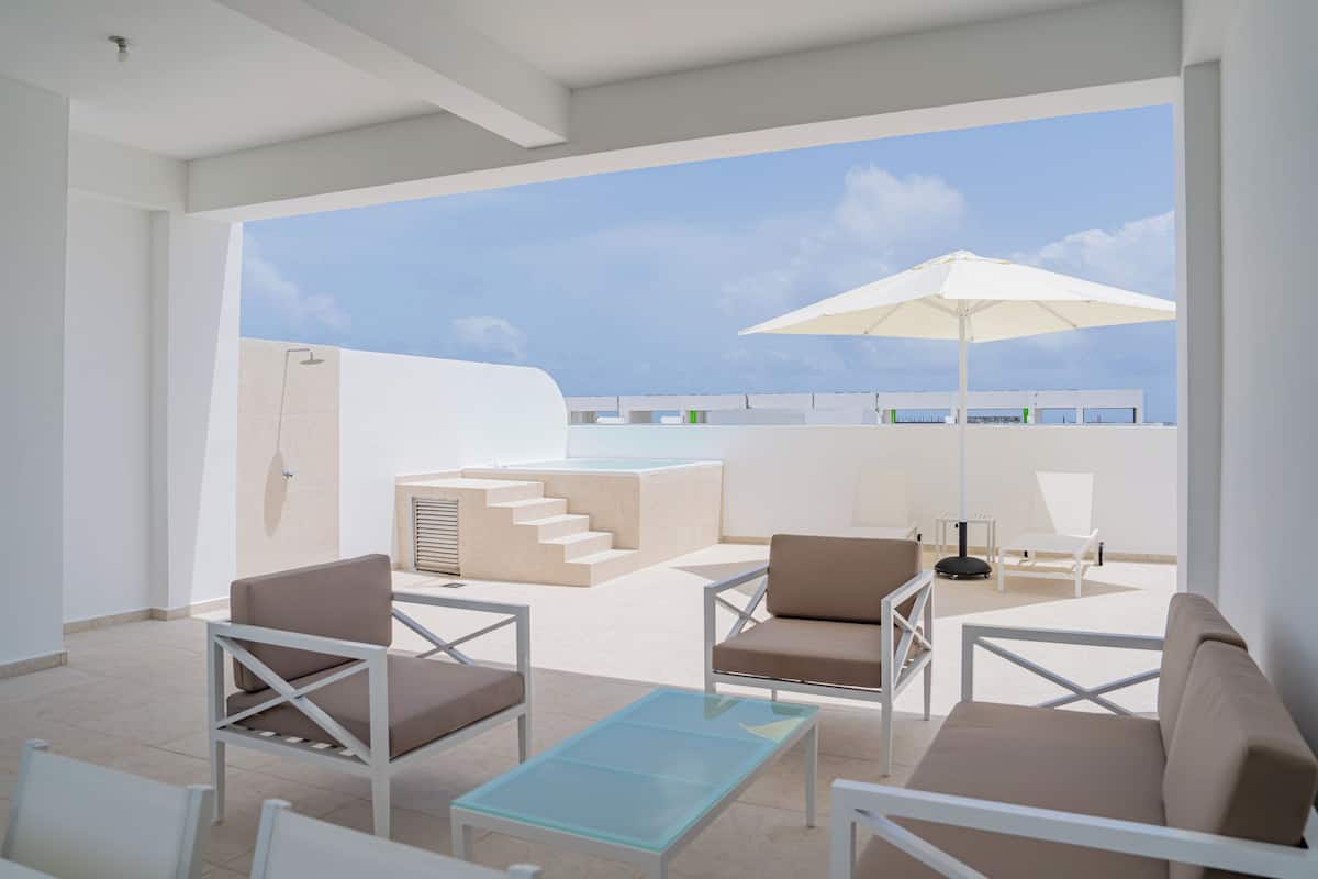 A modern outdoor lounge area with seating and a glass table, overlooking a pool and a clear sky.