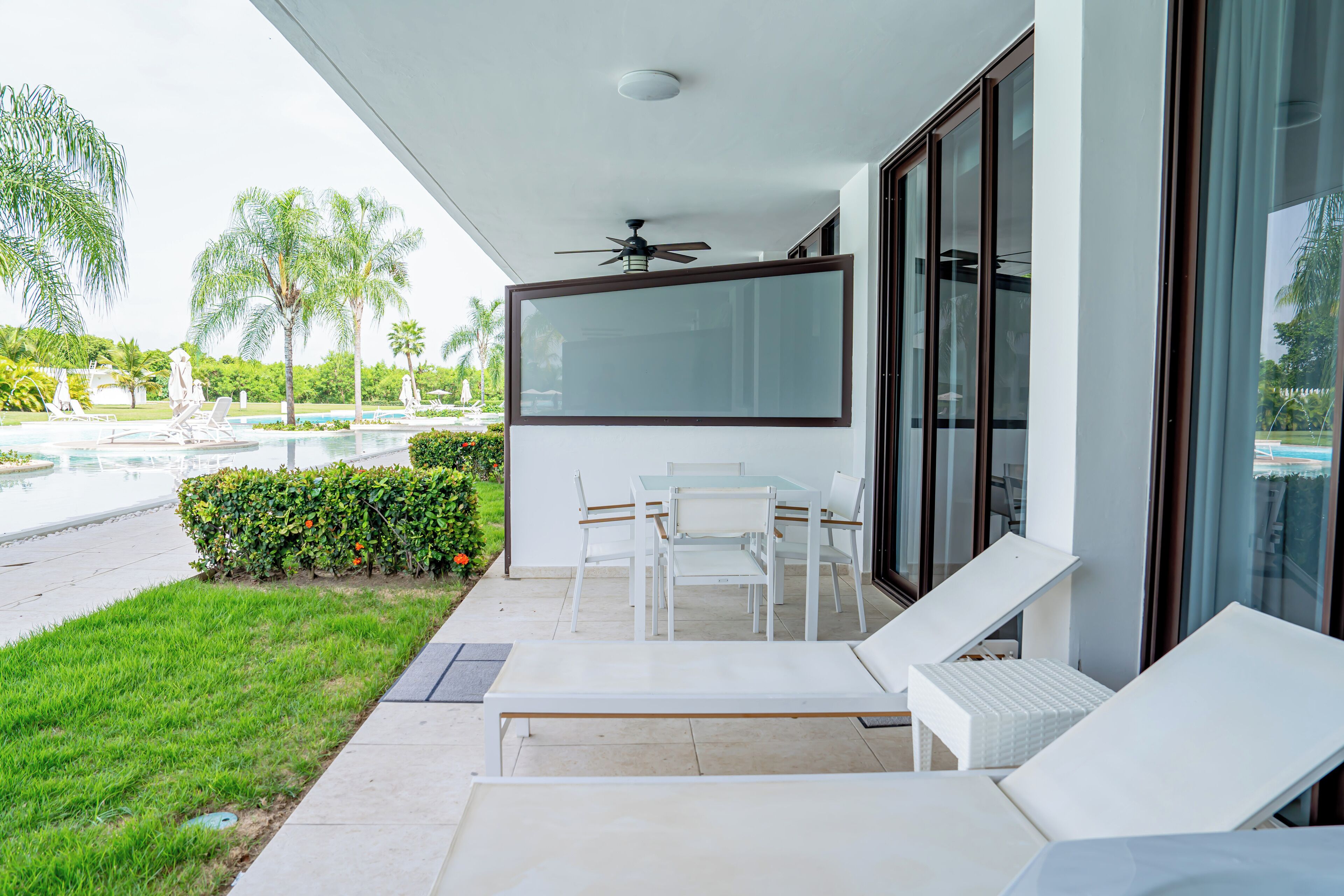 Un área de estar exterior moderna con sillones blancos y una mesa, con vista a una piscina y abundante vegetación.