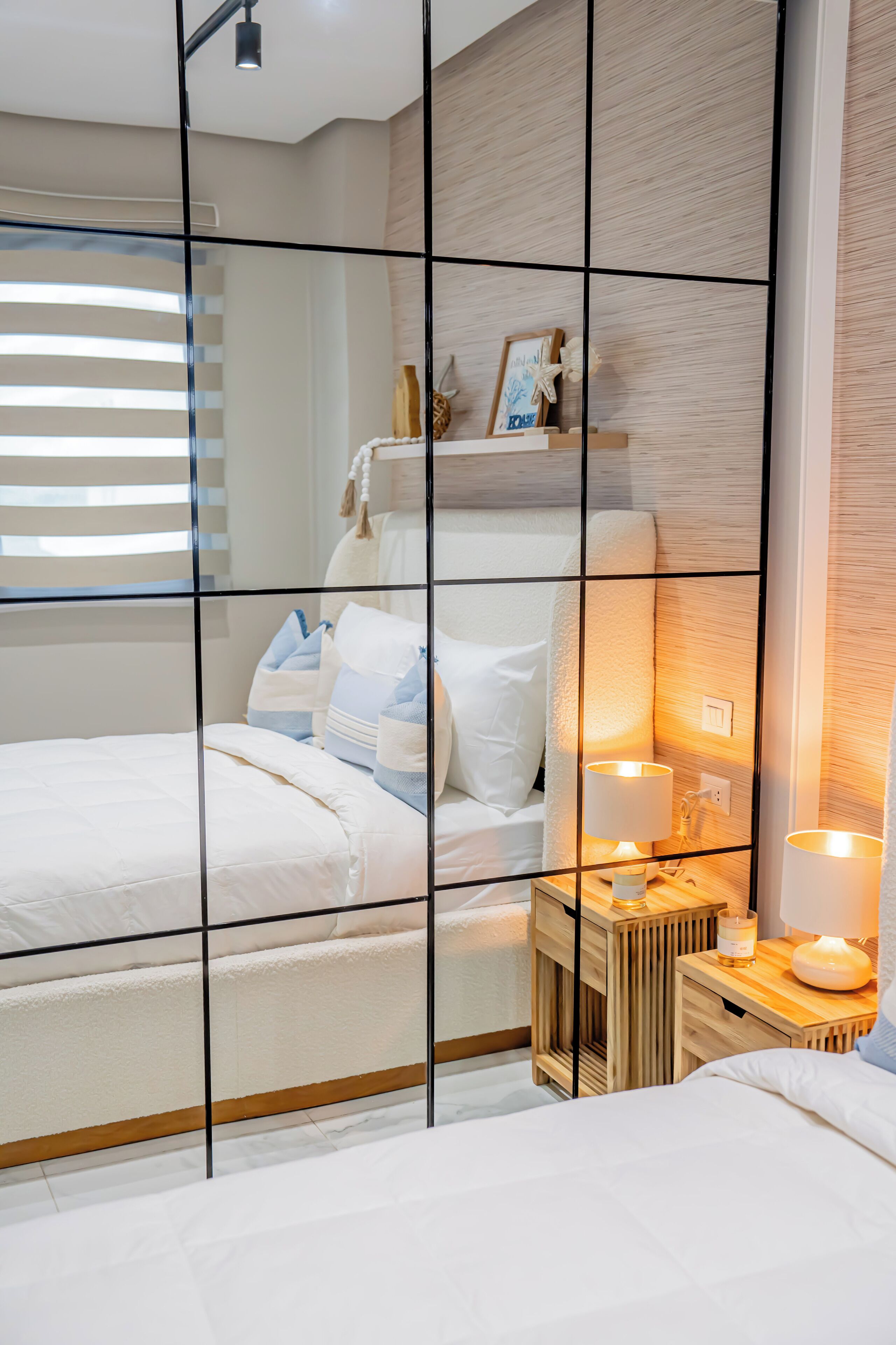 A modern bedroom with a large bed, bedside lamps, a small wooden nightstand, and a window with blinds.
