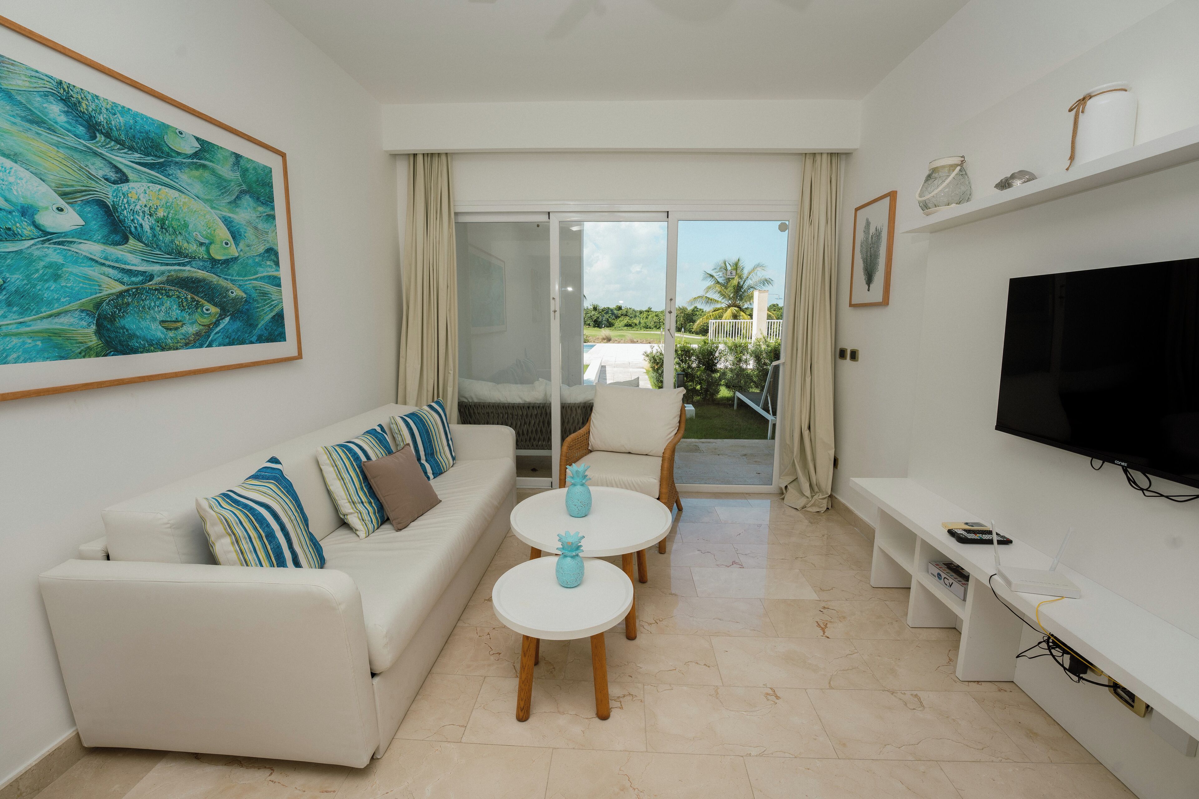 Un salon moderne avec un canapé blanc, deux tables rondes, un téléviseur à écran plat et un balcon offrant une vue sur la verdure.