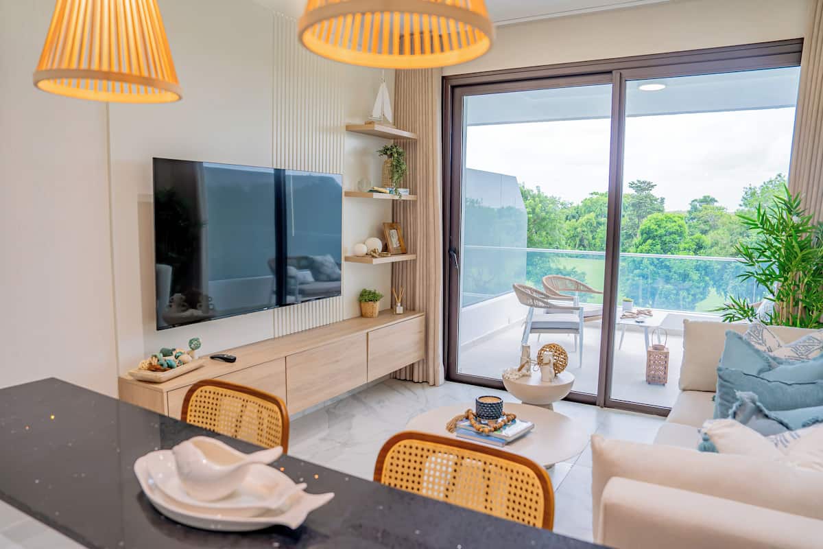 A modern living room with a flat-screen TV, wooden shelves, a dining area with a table and chairs, and a balcony with a view of greenery.