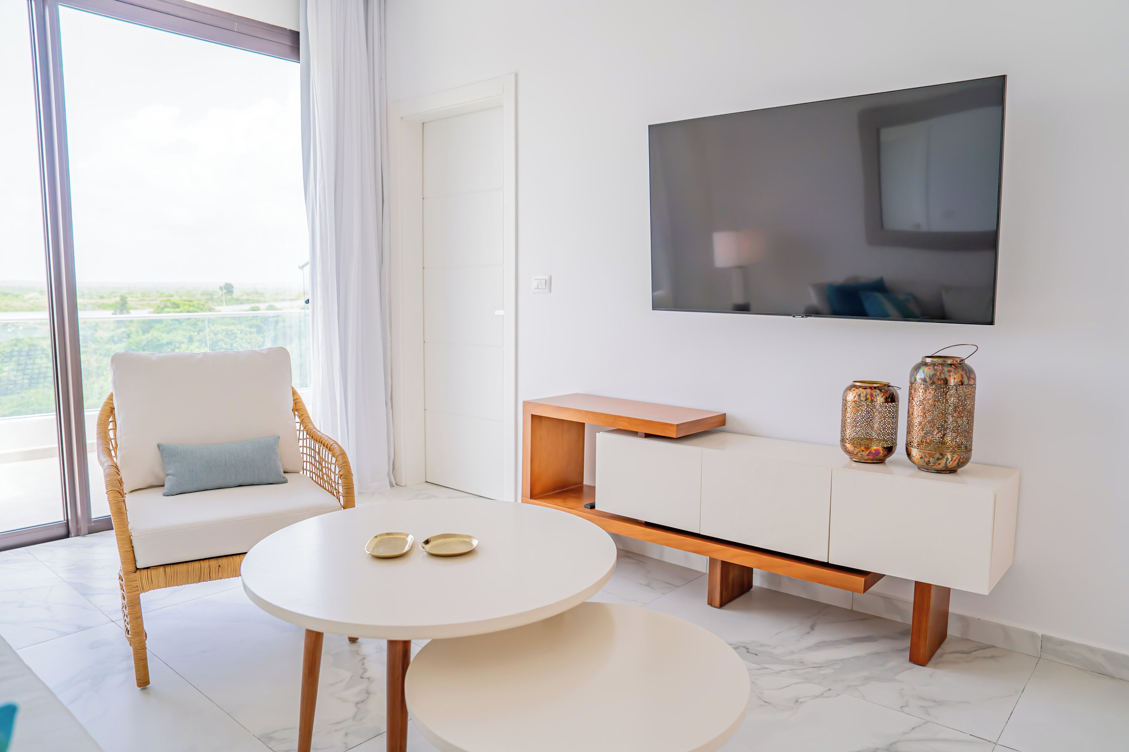 A modern living room with a white sofa, a round table with two plates, a TV mounted on the wall, and a wooden cabinet with decorative vases.
