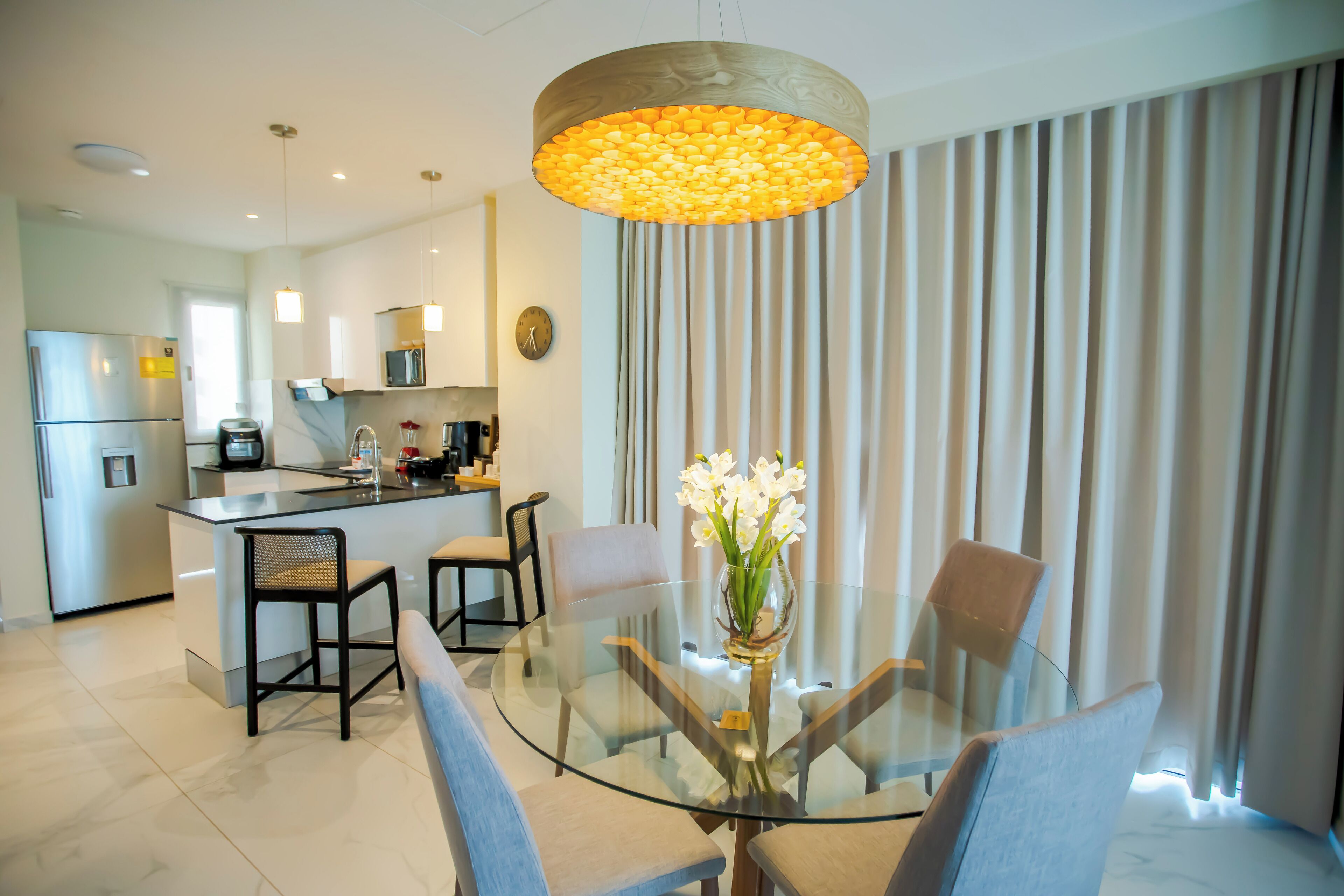 A modern kitchen with a central island, a dining area with a glass table and chairs, and a large window with curtains.