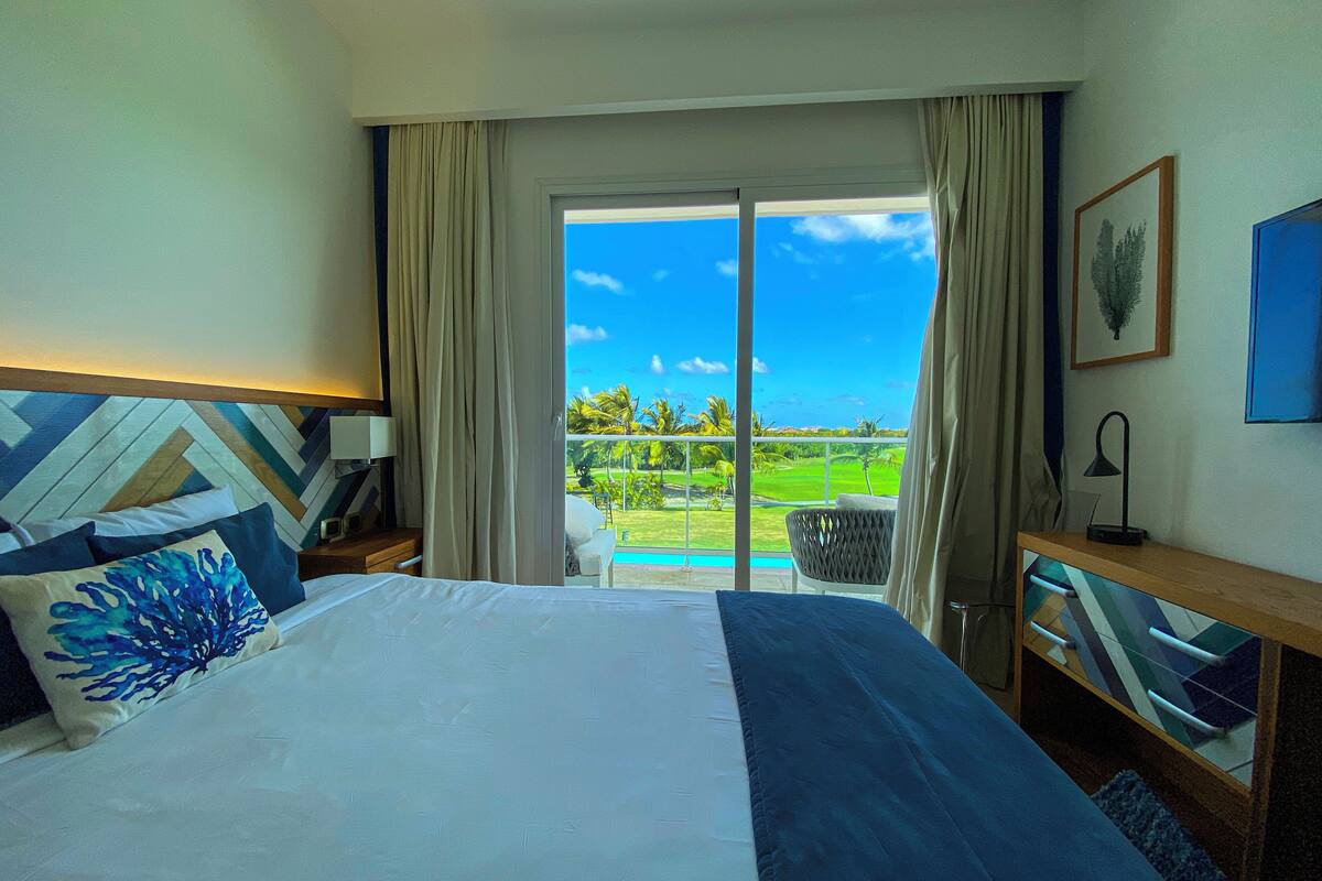 A hotel room with a bed, a nightstand, a desk, and a view of a golf course through a sliding glass door.