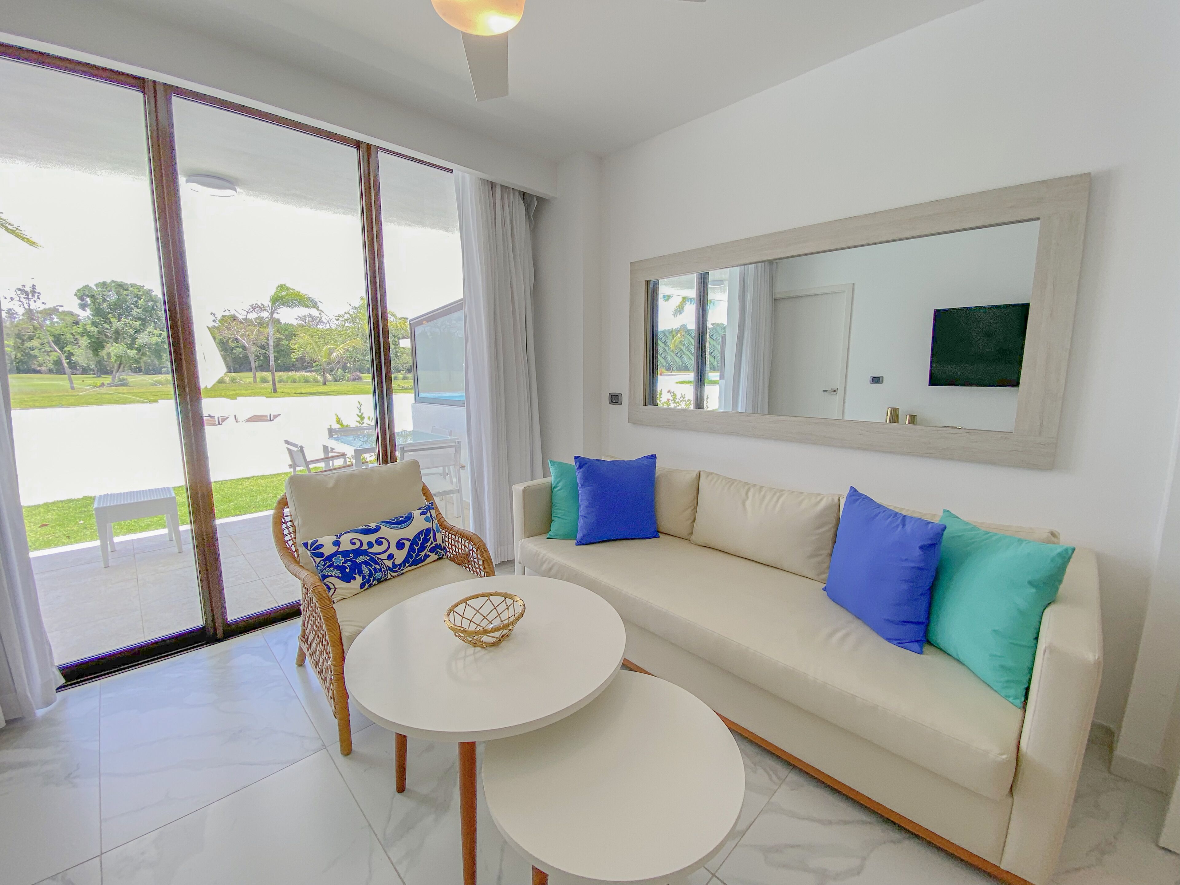 Un salon moderne comprenant un canapé, deux tables rondes, un fauteuil en osier et une grande fenêtre donnant sur une piscine et un espace verdoyant.