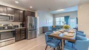 A modern kitchen with stainless steel appliances, a dining area with blue chairs, and a living room with a sofa and a TV. - 4012 Vista Cay (Orlando)