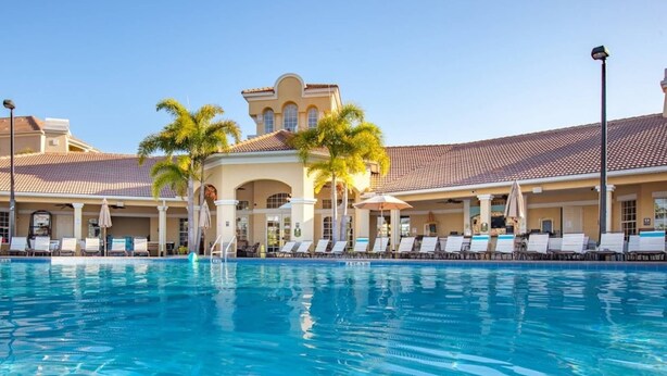 A swimming pool with lounge chairs and umbrellas, surrounded by a building with a tiled roof and palm trees. - 4012 Vista Cay (Orlando)