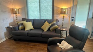 A living room with a dark grey sofa, two side tables with lamps, a wooden floor, and a door with blinds. - Moonlight on the White- Fayetteville river cabin (Springdale)