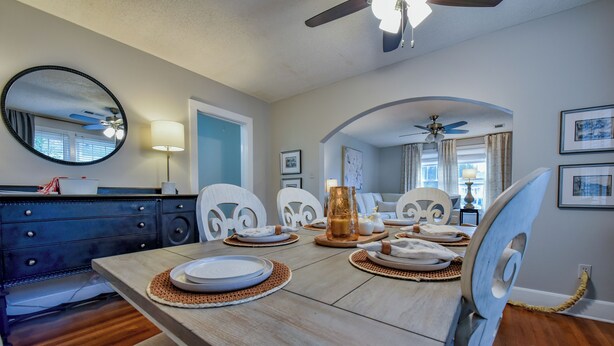 A dining area with a wooden table set for four, white chairs, a ceiling fan, and a large mirror on the wall. - Bama Bungalow - Book for football season and more! (Tuscaloosa)