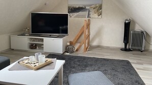 Quarto de sótão acolhedor e bem iluminado, com televisão de tela plana, um quadro de uma praia, um radiador e uma escada de madeira.