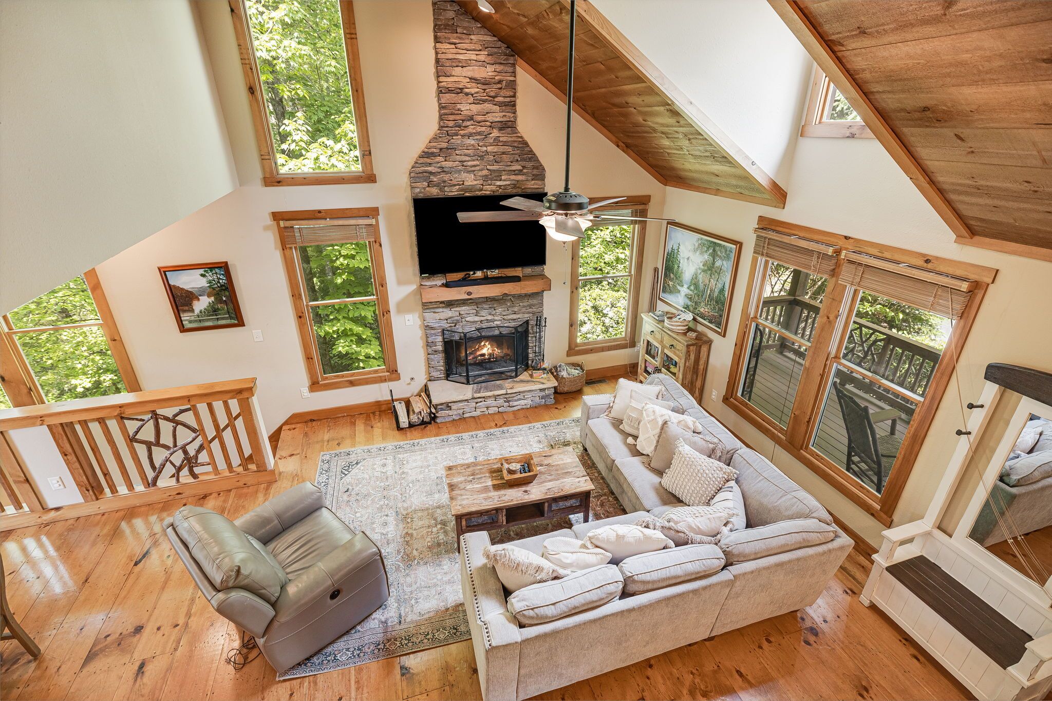 A spacious living room with a stone fireplace, wooden ceiling, and large windows offering views of the outdoors.