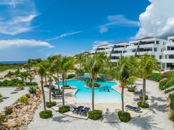 Um resort com piscina central rodeada por palmeiras e espreguiçadeiras, com um edifício moderno ao fundo.
