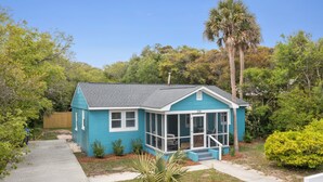 Einbungalow mit blauer Fassade, einer überdachten Veranda und einem kleinen Vorgarten mit einer Palme und Büschen.