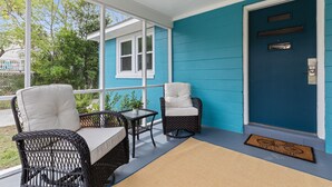 Eine Veranda mit zwei Korbstühlen und einem kleinen Tisch, eine blaue Tür mit Fußmatte und Blick auf ein Haus durch große Fenster.