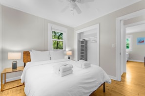 A bedroom with a large bed, a nightstand with a lamp, a window with blinds, and a door leading to another room.