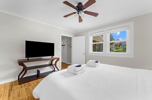 A hotel room with a bed, a television on a wooden stand, a ceiling fan, a window with a view of buildings, and a door.