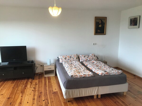Un dormitorio con cama, televisor, piso de madera, mesita de noche y lámpara colgante.