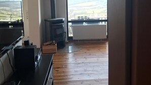 A living room with a TV, a wood-burning stove, and a large window with a view of the countryside. - Panoramic view of the Rhön (Bischofsheim an der Rhön)