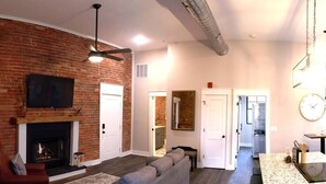 A living room with a fireplace, a sofa, and a television. There is a kitchen area visible through an open door. - The Double Diamond Loft (Boyne City)
