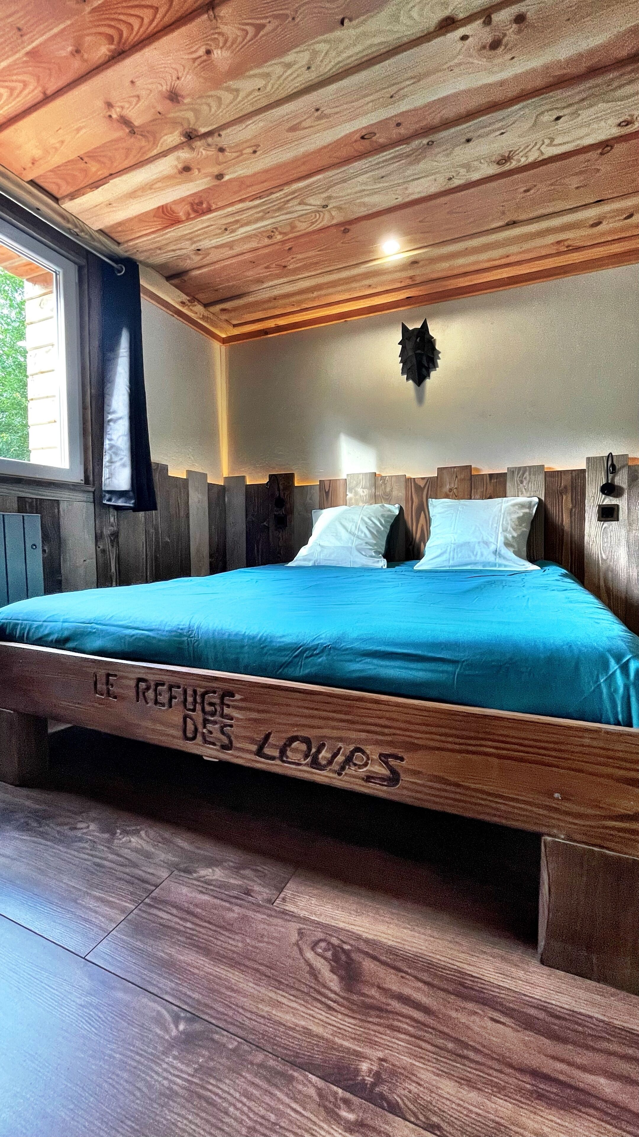 Ein hölzernes Bett mit blauer Matratze und Kissen, ein Holz-Kopfteil und ein Fenster mit Vorhang.