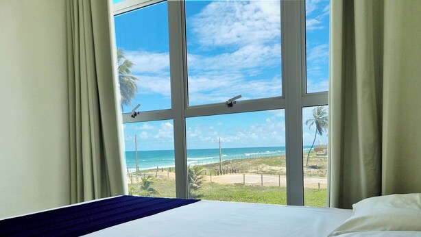 A hotel room with a large window offering a view of the beach and ocean. - Apê p/ 7 Pessoas: Vista Mar, Piscina e Garagem (Barra de São Miguel)