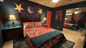 A bedroom with a large bed, two bedside tables with lamps, a ceiling fan, a tapestry with a moon and stars, a mirror, and a red door. - Pioneertown Bohemian Getaway! (Pioneertown)