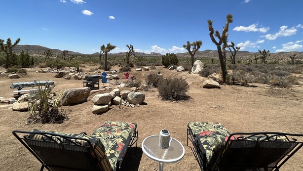 An outdoor seating area with two lounge chairs and a small table, surrounded by desert landscape and Joshua trees. - Pioneertown Bohemian Getaway! (Pioneertown)