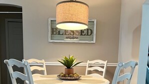 A dining area with a round table, white chairs, a hanging light fixture, and a framed sign on the wall.