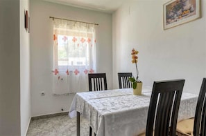 Un coin repas avec une table recouverte d’une nappe blanche, deux chaises, une orchidée en pot et une fenêtre ornée de rideaux à motifs floraux.