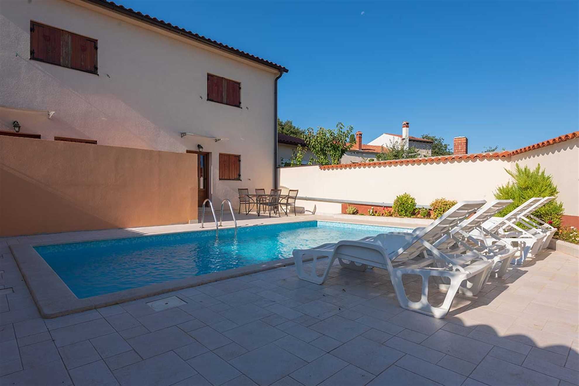 Una piscina con sillas de descanso, un patio empedrado y un edificio con fachada blanca y contraventanas rojas.