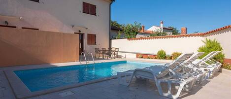Una piscina con sillas de descanso, un patio empedrado y un edificio con fachada blanca y contraventanas rojas.