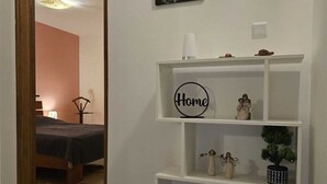 A hallway with a white console table displaying a 'Home' sign, a small potted plant, and a black mug. A door leads to a bedroom with a bed and a nightstand.