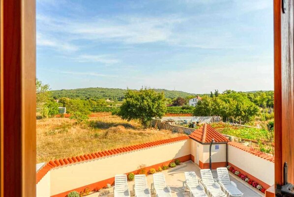 Un balcón con sillones blancos, vista a un paisaje verde y un edificio con tejado de tejas rojas.