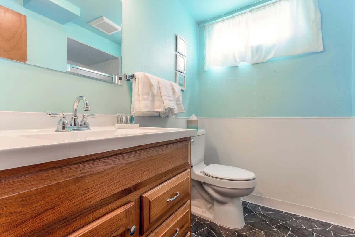 A bathroom with a wooden vanity, a white toilet, and a window with a white curtain.